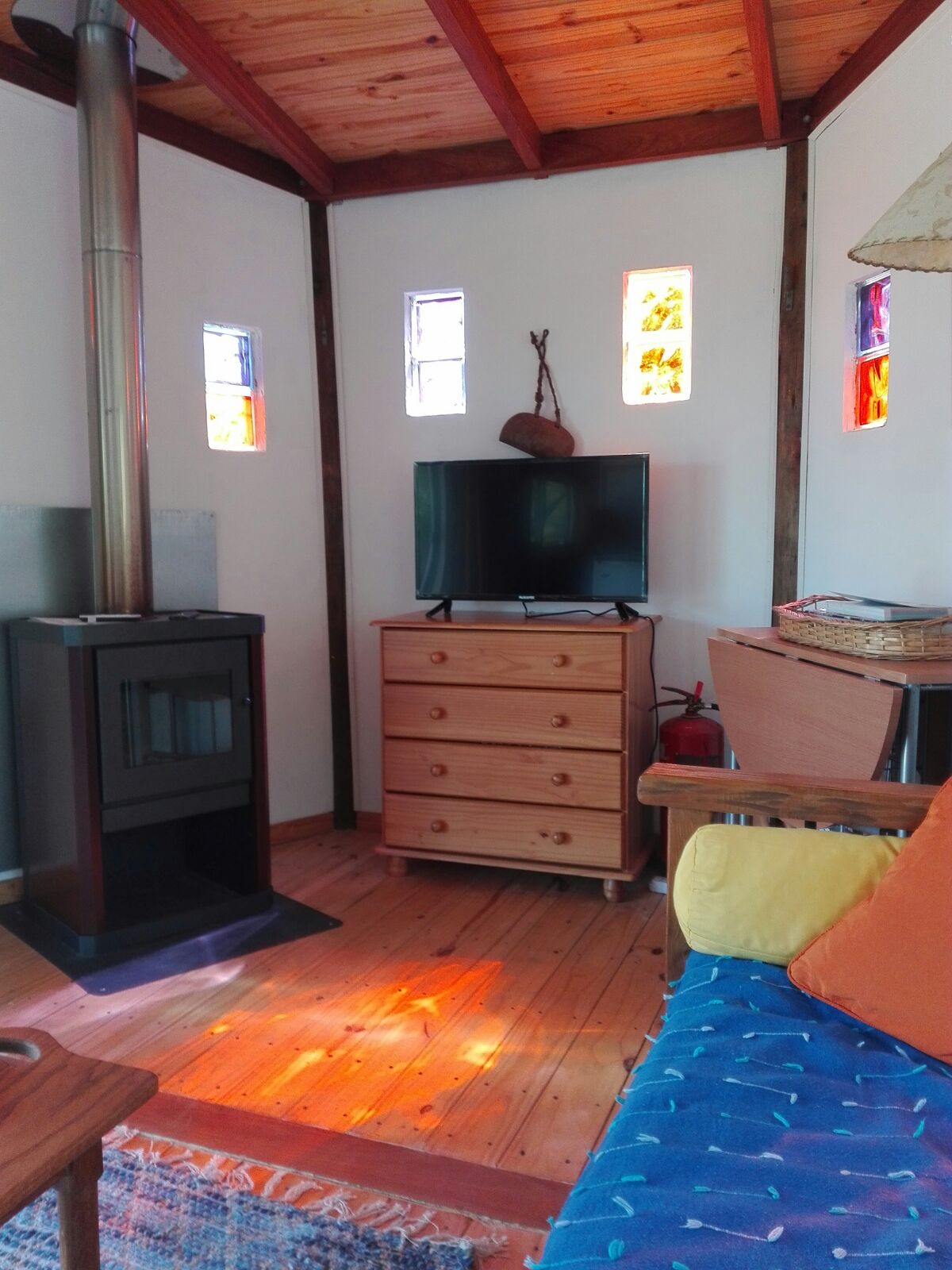 A cozy living area is displayed, featuring a wooden dresser and a television. Stained glass windows allow colorful light to filter in, adding warmth to the space. A comfortable sofa is complemented by a rustic lamp, with light reflecting on the wooden floor.