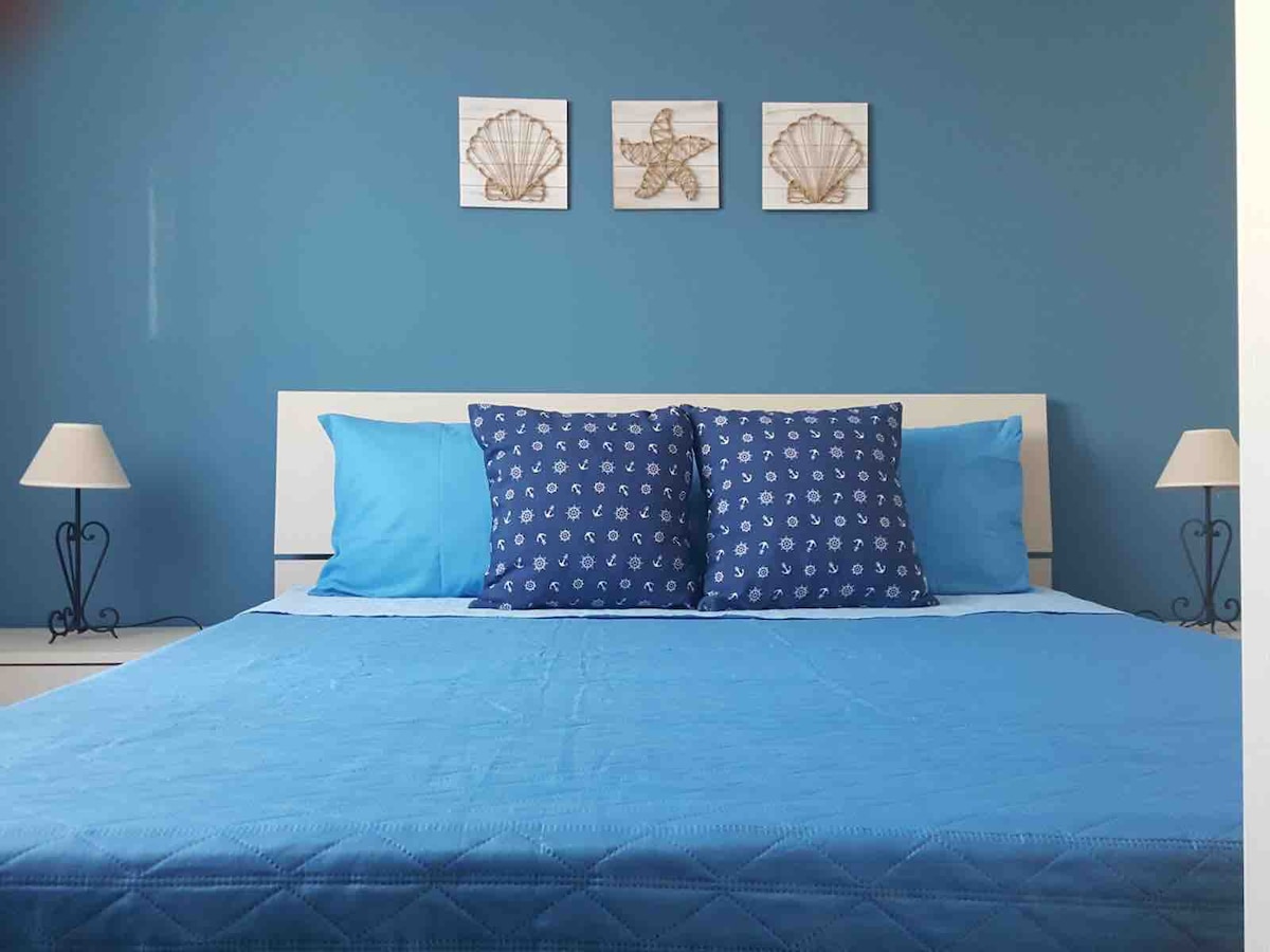 A calming bedroom features a blue wall and a spacious bed dressed in blue linens. Two decorative pillows with nautical patterns are positioned at the head of the bed. On either side, slender lamps provide additional lighting, complementing the overall serene ambiance.