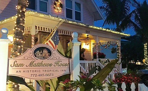 The exterior of the Sam Matthews House is adorned with festive lights, creating a welcoming atmosphere. A prominent sign displays the name of the historic inn. Lush greenery and palm trees surround the entrance, enhancing the tropical setting.
