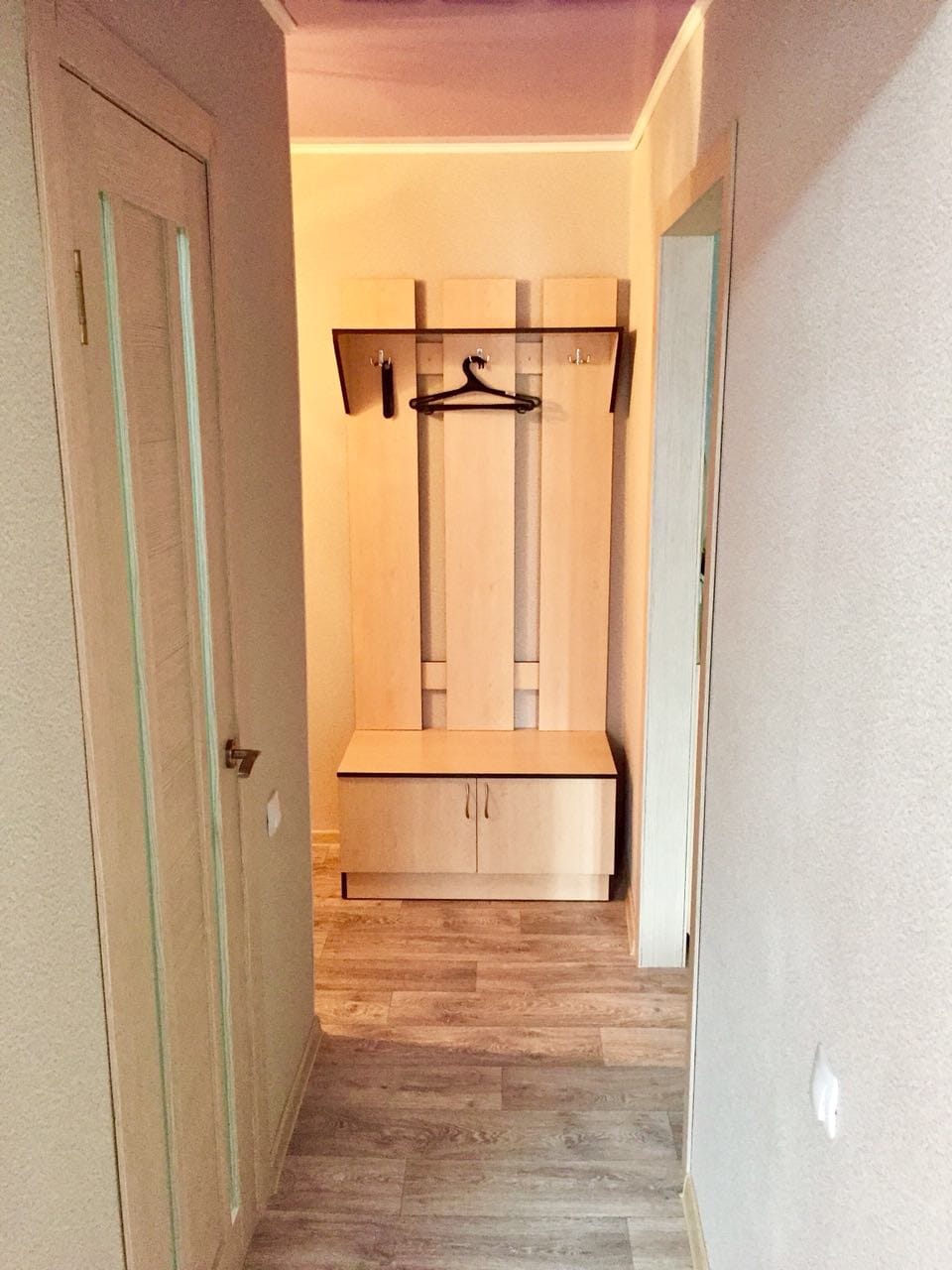 A well-organized entryway features light wooden paneling and a neatly arranged coat rack with a hanger. A compact bench with storage space is positioned below, providing functionality and ease for guests. The soft lighting contributes to a welcoming atmosphere.