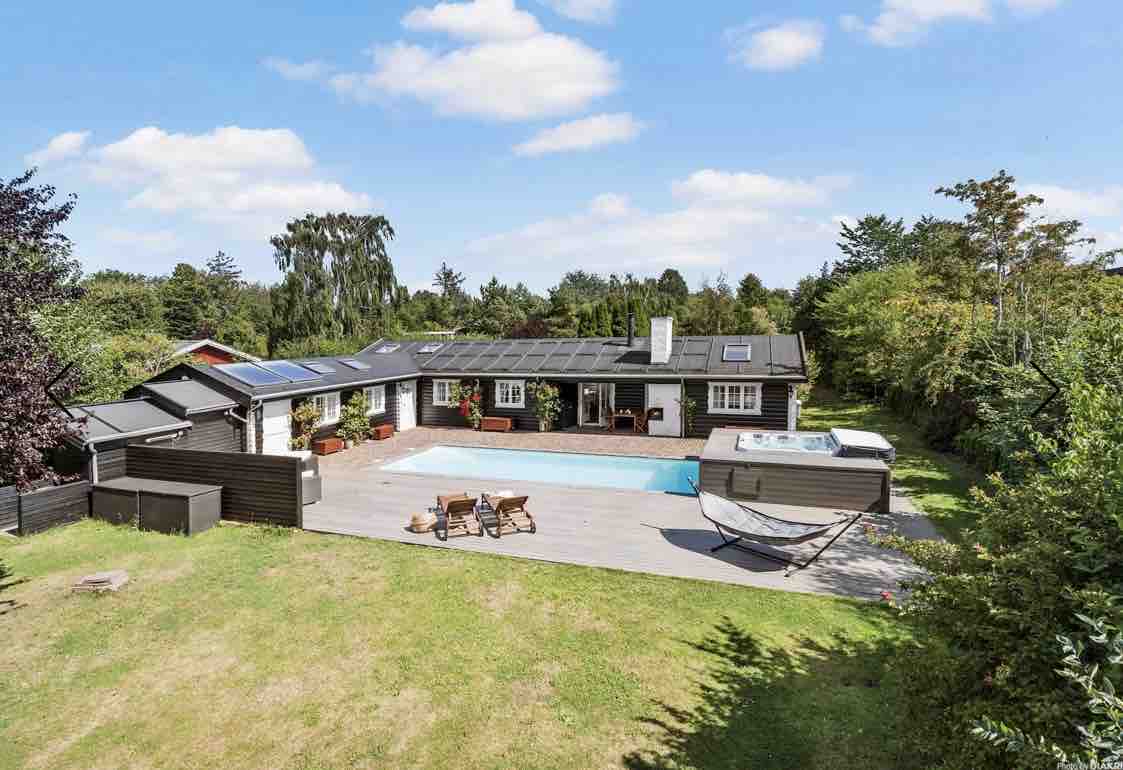 A spacious outdoor area features a well-maintained garden surrounding the property. A swimming pool is centrally located, accompanied by a hot tub. Lounge chairs are positioned nearby, while a hammock adds a relaxing touch. Surrounding trees enhance the sense of privacy and tranquility.