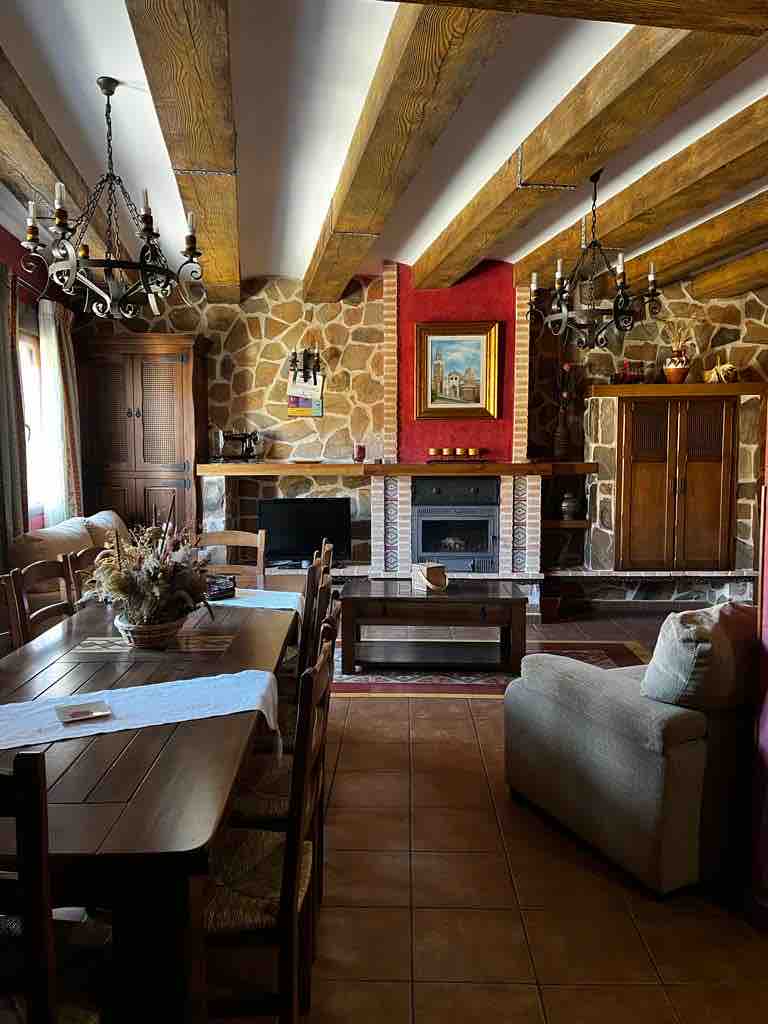 An inviting living area features a large wooden dining table surrounded by chairs, complemented by rustic stone walls and exposed wooden beams. A cozy seating arrangement is positioned near a classic fireplace, enhancing the warm ambiance. Natural light filters through the windows adorned with soft coverings.
