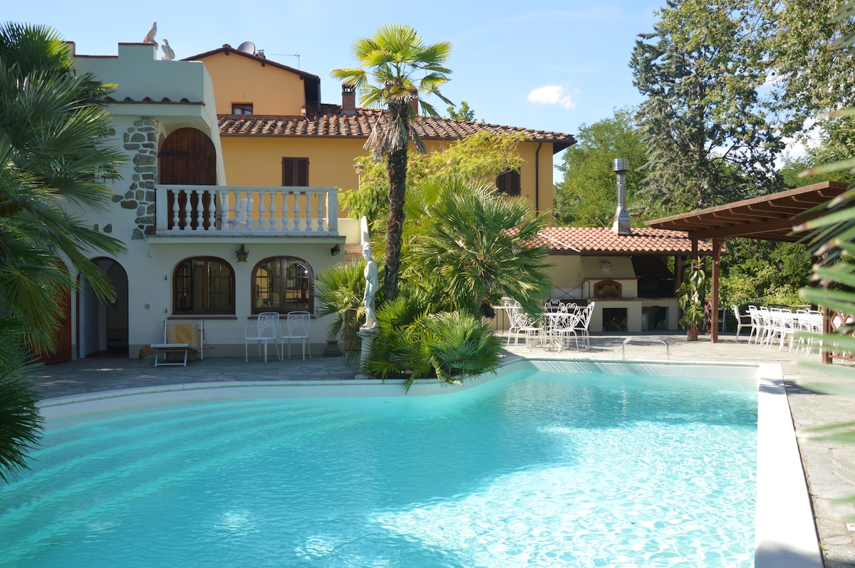 The outdoor area features a large swimming pool surrounded by lush greenery and palm trees. A terrace with outdoor seating is visible, along with a covered dining area ideal for gatherings. The building displays traditional Tuscan architecture with arched windows and a rustic roof.