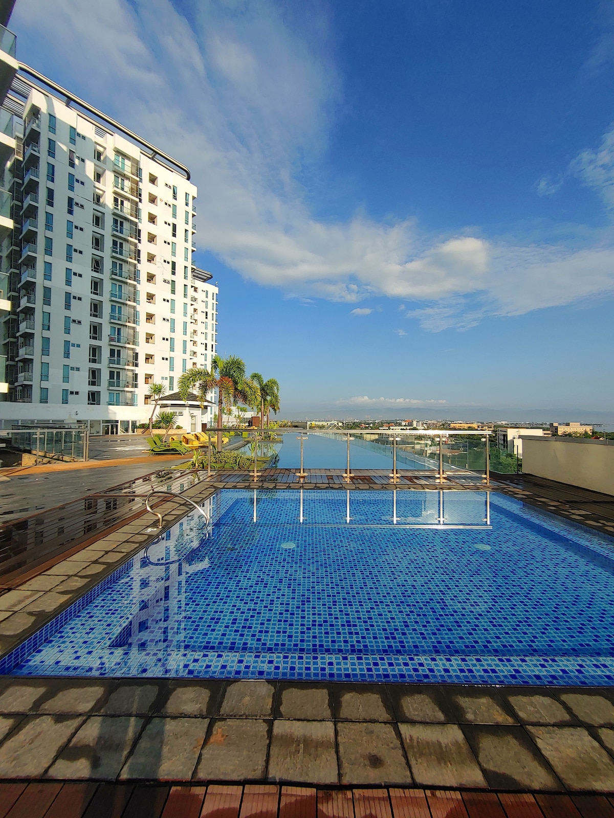 A serene outdoor pool area features a mosaic-tiled surface reflecting the sky. Lush palm trees line the edges, and a modern building towers in the background. The water glistens under sunlight, contributing to the tranquil ambiance of the space.
