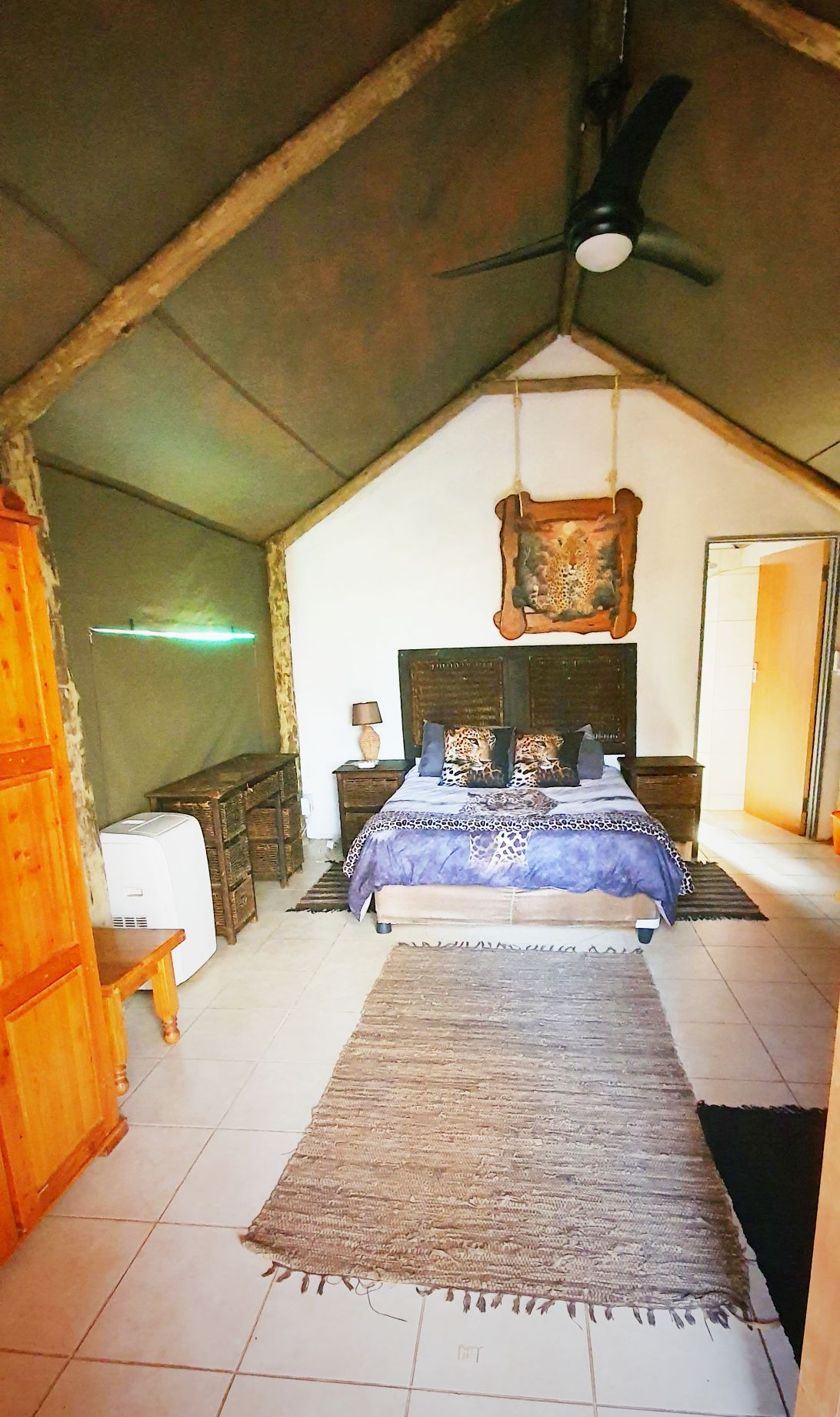 A spacious sleeping area features a double bed with decorative blue bedding and an artistic wall hanging above the headboard. Natural materials are evident in the wooden furniture, while a ceiling fan provides ventilation. A textured rug adds warmth to the tile floor, with soft lighting enhancing the ambiance.