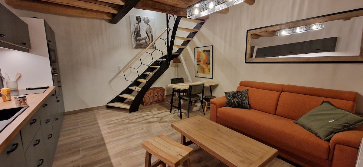 An open living space is displayed, featuring a warm-colored sofa with a decorative cushion. A dining area consists of a table and four chairs. A wooden staircase leads to an upper level, with a wall art piece and recessed lighting accentuating the area.