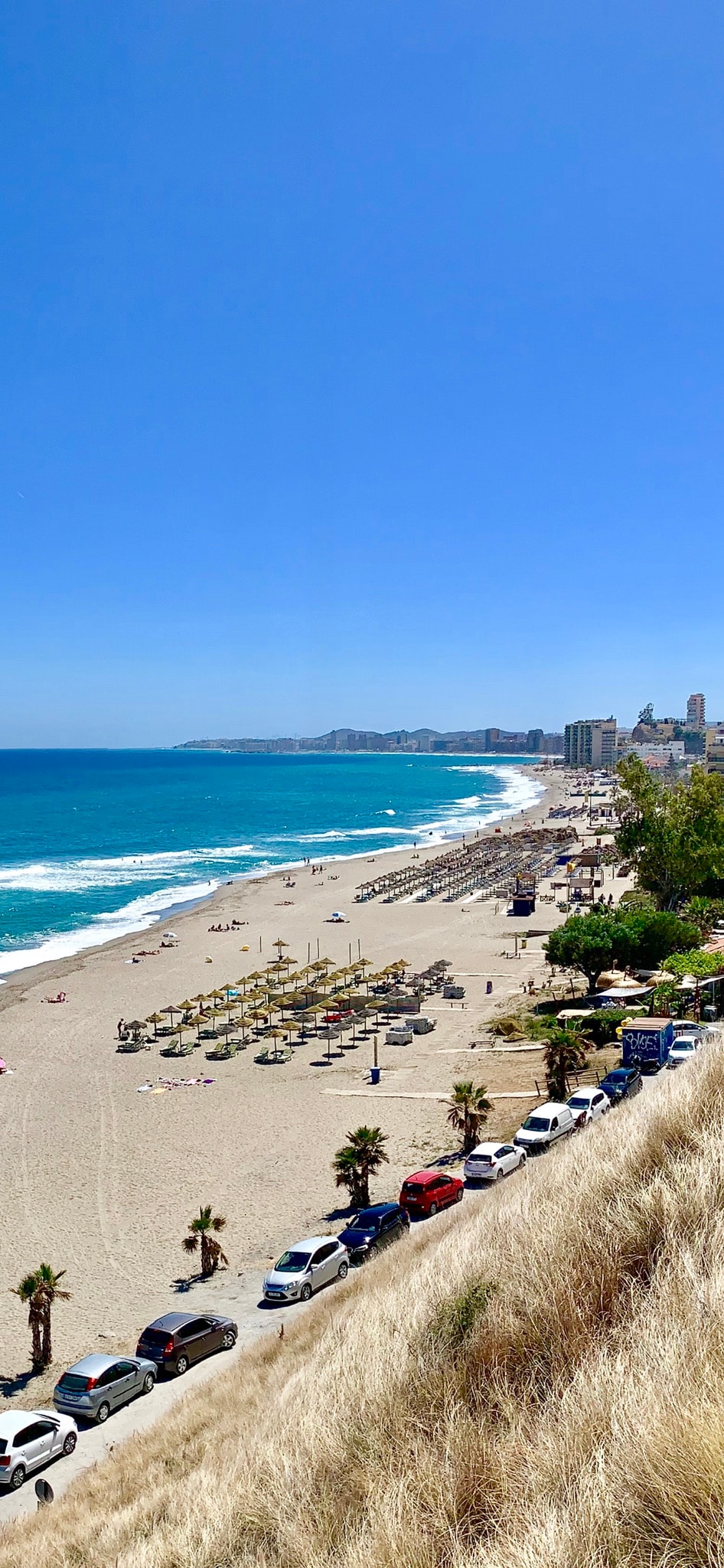 An expansive beach scene stretches along the coastline, featuring golden sand and gentle waves. Sun loungers and umbrellas are arranged near the shoreline, while palm trees provide natural greenery. A line of cars is parked along a road overlooking the beach, set against a clear blue sky.