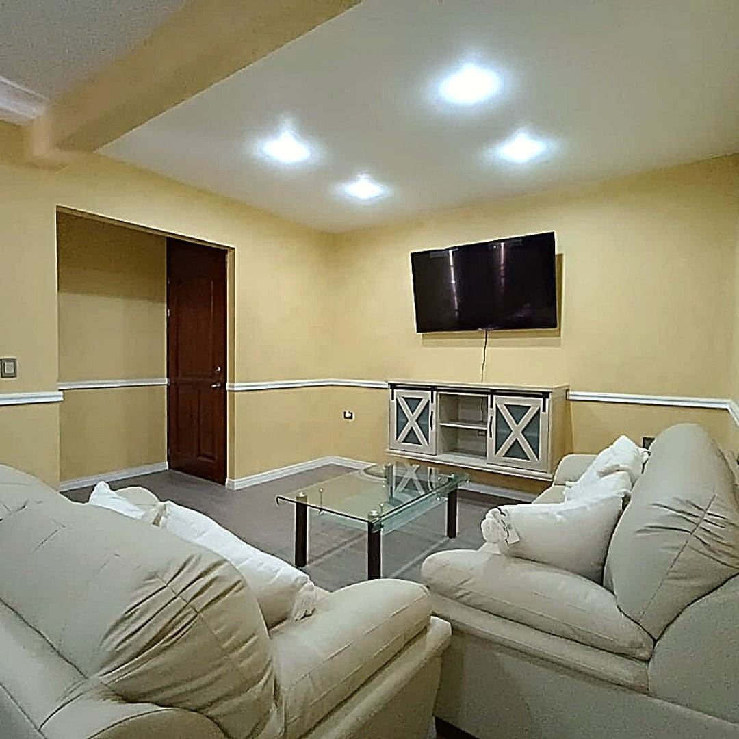 A cozy living area features light-colored sofas surrounding a glass coffee table. A mounted television is positioned on a warm yellow wall, while soft lighting enhances the relaxed ambiance. A doorway leads to another space, adding to the room's openness.