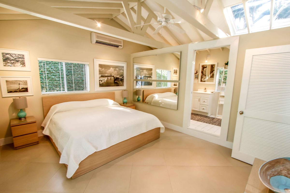 A spacious bedroom is showcased, featuring a wooden bed frame with a light-colored bedspread. Natural light filters in through multiple windows. Photographic artwork adorns the walls, and a mirror reflects the en-suite bathroom area, which is visible beyond an open door.