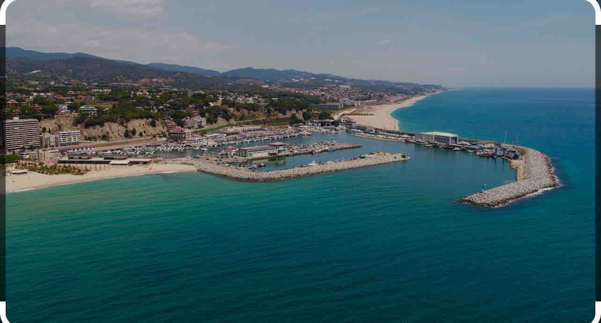 An aerial view captures the coastline, showcasing a serene beach and a marina lined with boats. The clear blue water contrasts with the sandy shore, while green hills rise in the background, creating a tranquil setting near the village.