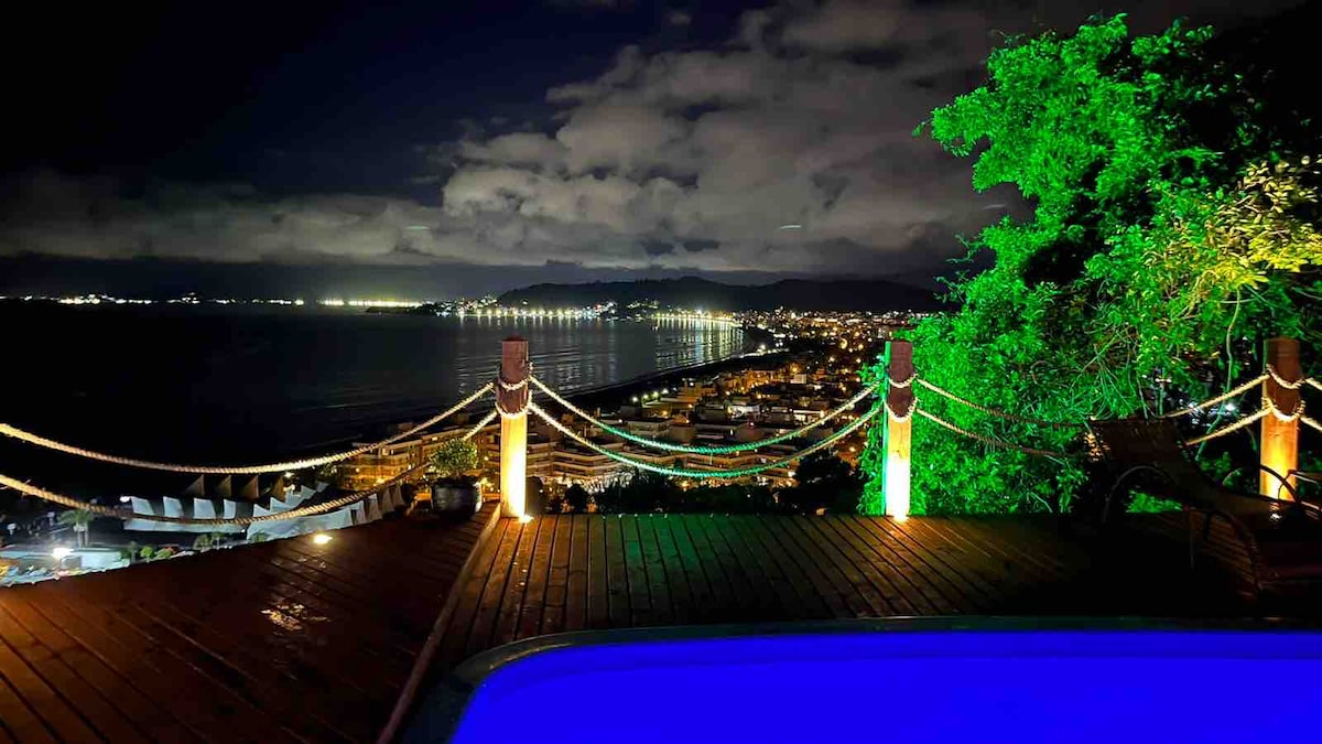 The pool area is illuminated by soft lighting, reflecting a serene blue hue. A panoramic view of the coastline is visible, with city lights twinkling along the shoreline. Lush greenery frames the scene, enhancing the peaceful ambiance of the outdoor space.