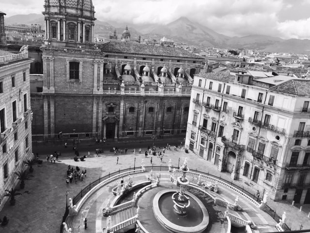 A panoramic view captures a historic square featuring a central fountain and surrounding architectural details. Notable structures include a prominent church and elegant facades, framed by mountain silhouettes in the background. People can be seen leisurely enjoying the space, highlighting the vibrant atmosphere.