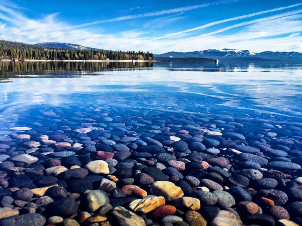 A clear lake is seen with a vibrant display of smooth, multicolored stones visible beneath the water's surface. The calm waters reflect the blue sky and wispy clouds, creating a serene atmosphere with distant mountains framing the horizon.