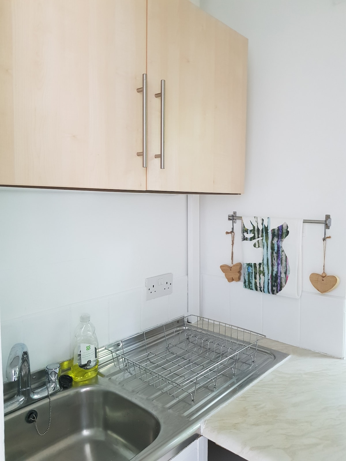 The kitchenette showcases light wood cabinets and a stainless steel sink. A metal drying rack rests on the counter beside a bottle of dish soap. A colorful towel hangs from the wall, adorned with decorative items in the shape of hearts.