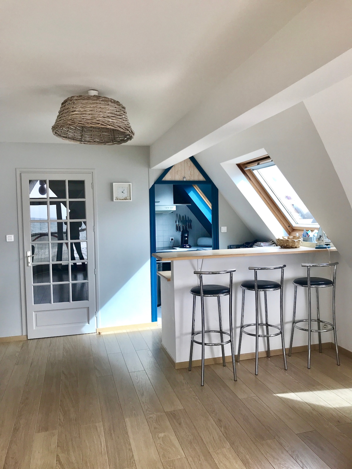 A modern kitchen area is illuminated by natural light streaming through a slanted window. A small bar with three metal stools is positioned beside an open kitchen with blue cabinetry. Light wooden flooring enhances the airy feel of the space.