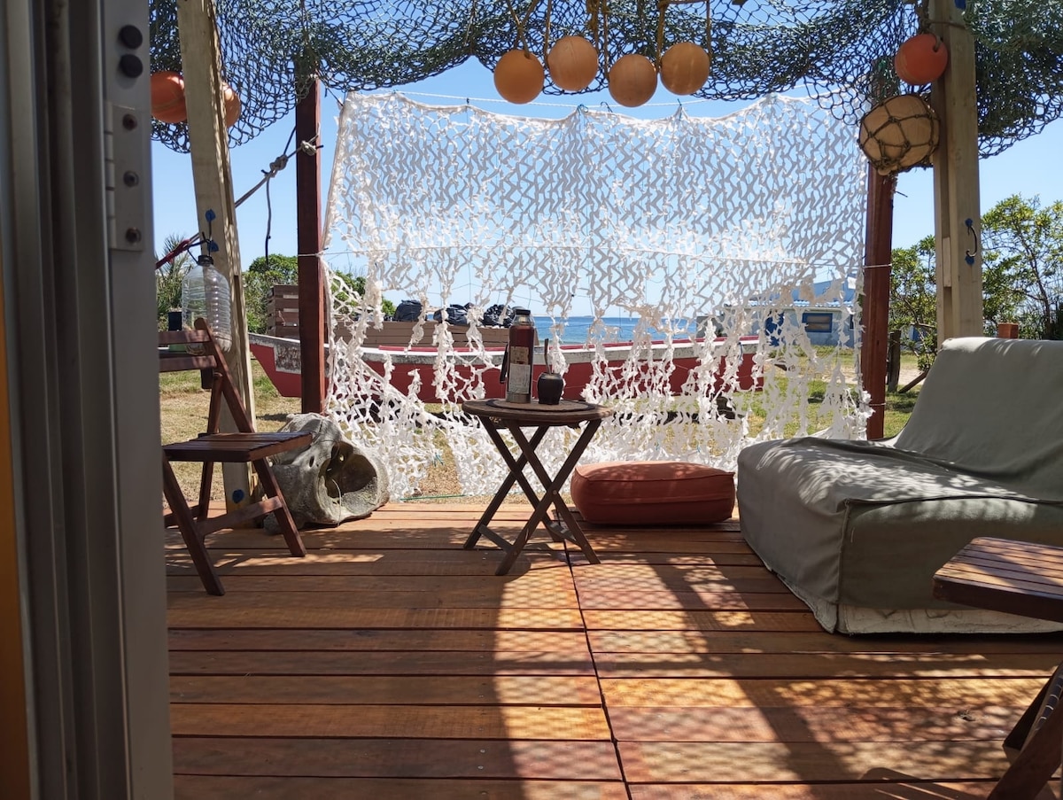 A deck area features wooden flooring with a small table and two chairs positioned for relaxation. A cozy seating arrangement includes cushions, surrounded by a decorative net and providing a view of the ocean. Natural light filters in, enhancing the serene atmosphere.