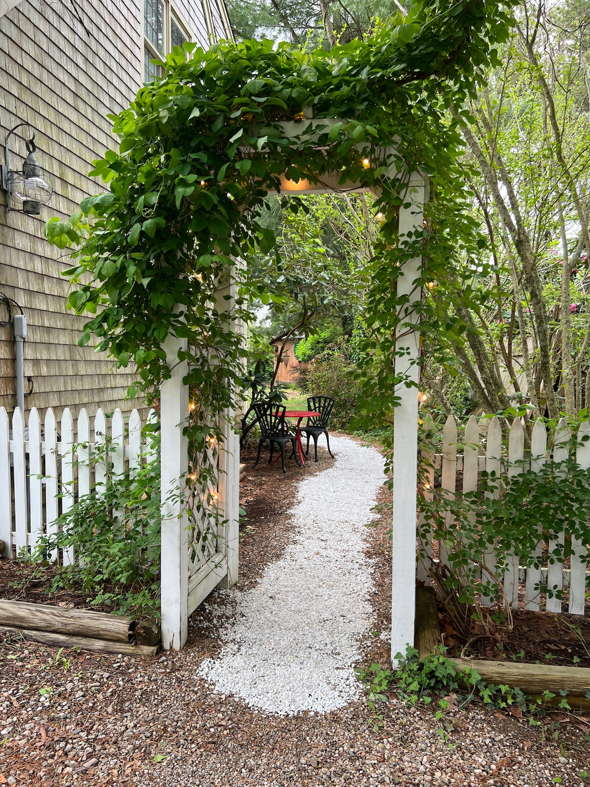 A white wooden arch covered in greenery leads to a gravel path. On one side, a small patio area with a black table and chairs is visible, surrounded by lush foliage. The setting captures a serene outdoor space within a well-maintained garden.