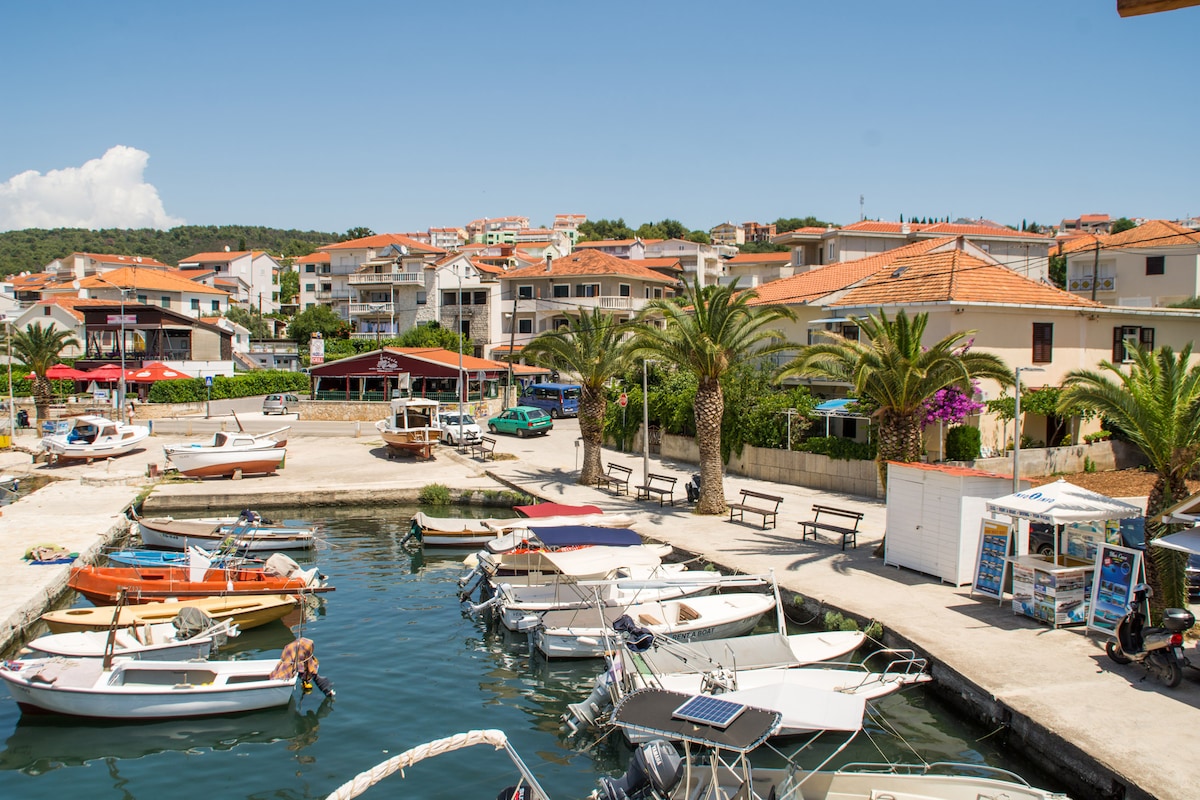 A scenic marina is visible, featuring several boats anchored along the dock. Lush palm trees line the waterfront, and a quaint village is seen in the background, showcasing buildings with terracotta roofs. Bright blue skies enhance the coastal atmosphere.
