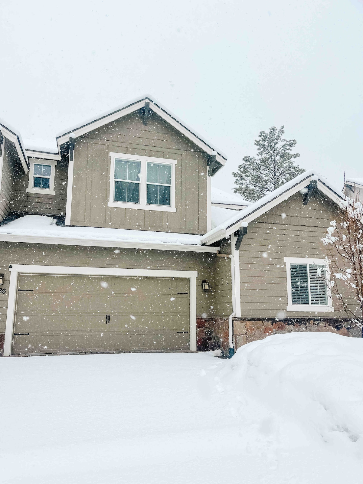 A two-story townhouse is shown blanketed in fresh snow, with a lightly colored exterior and a garage door visible. Flakes fall softly, creating a serene winter scene. Snow accumulates on the ground and around the entryway, highlighting the home's inviting facade.