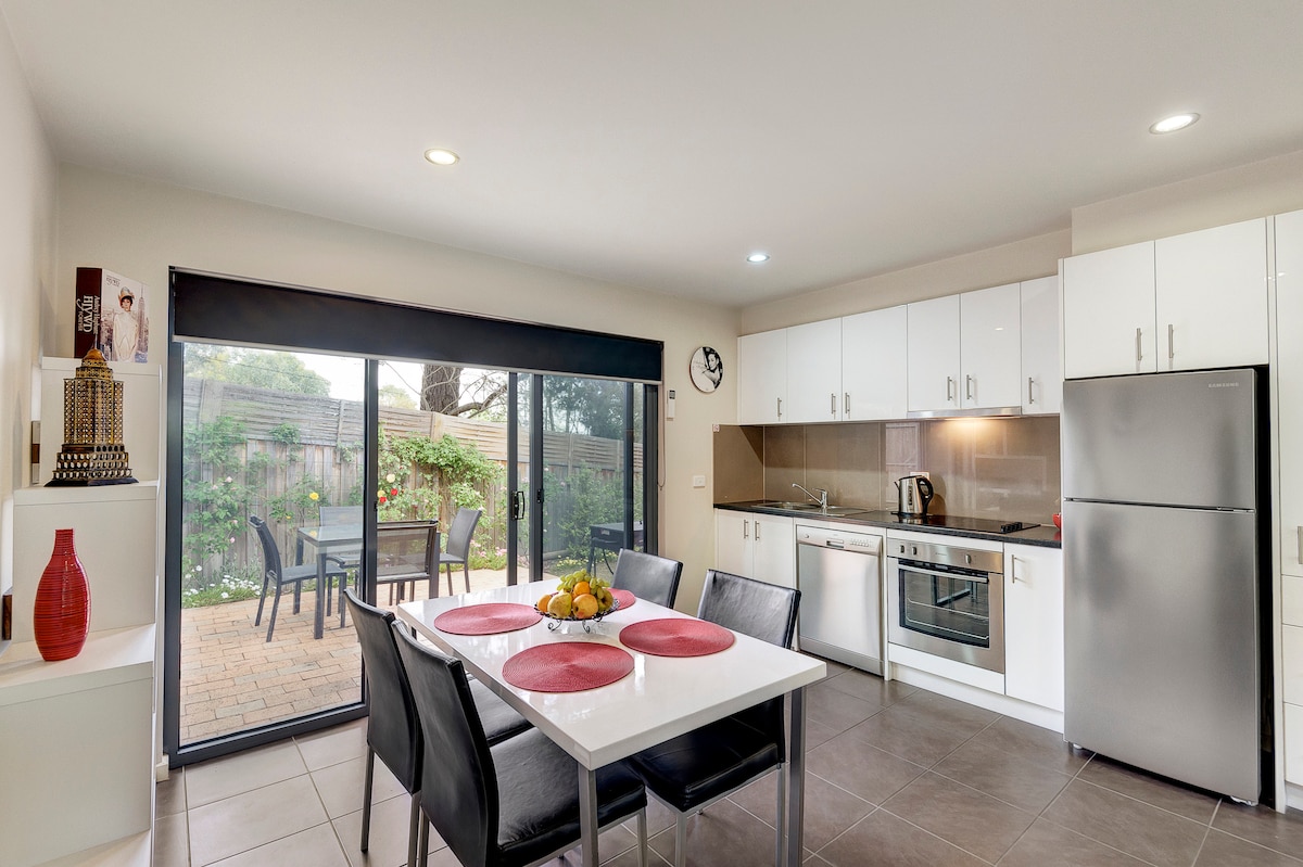 A modern kitchen and dining area is showcased with white cabinets and stainless steel appliances. A dining table with black chairs is set with red placemats. Large sliding doors open to a brick-paved outdoor space featuring lush greenery.