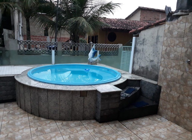 A round pool is set within a stone enclosure, featuring steps leading up to it. Surrounding greenery is visible, with a palm tree in the background. The area is paved, and a shower is positioned nearby, providing a refreshing outdoor experience.