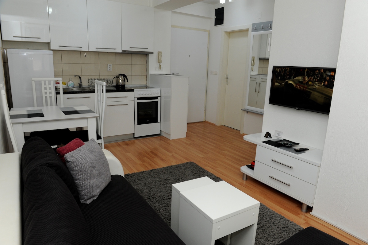 A modern kitchen area is visible, featuring sleek white cabinetry and appliances, including an oven and a dining table with four chairs. A comfortable corner sofa with pillows is positioned beside a small coffee table. A wall-mounted LED TV displays a scene from a movie.