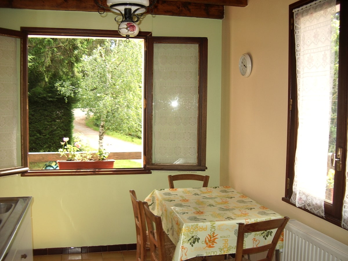 A kitchen area is presented with a wooden table surrounded by four chairs, set beneath a rustic light fixture. Natural light floods in through large windows, showcasing views of greenery outside. A clock is mounted on the wall, indicating the rural atmosphere of the space.