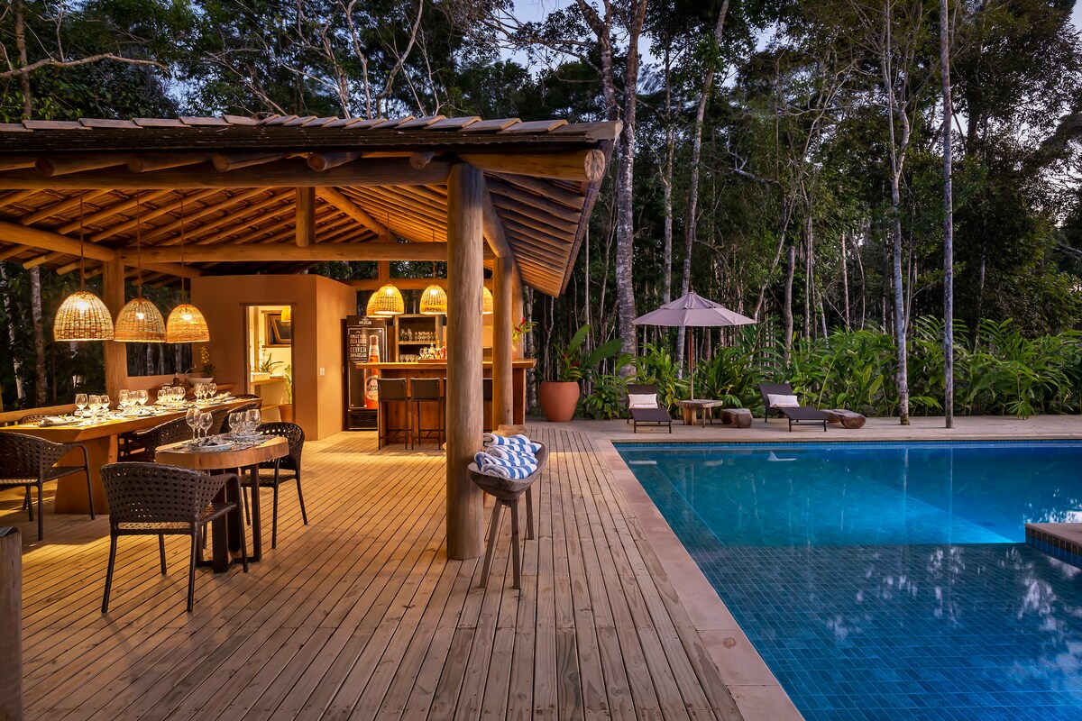 An outdoor area features a wooden deck surrounding a clear pool. A covered bar section is visible with pendant lights hanging overhead. Nearby dining tables are set with dishes. Lounge chairs sit along the poolside, surrounded by lush tropical greenery.