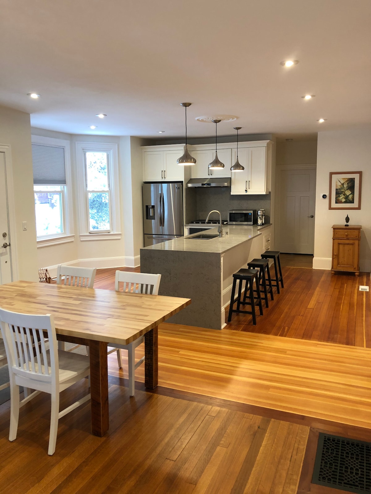 An open kitchen and living area are featured, showcasing modern appliances including a refrigerator and microwave. A wooden table with chairs is positioned prominently, while bar stools align with the kitchen island. Ample natural light enters through the windows, highlighting the hardwood floor.