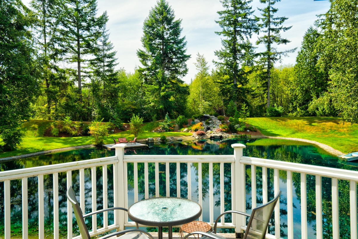 A private balcony is furnished with a glass-topped table and two chairs. The view includes a tranquil pond surrounded by lush greenery and trees, with a gently sloping bank leading to a well-maintained yard. Sunlight reflects off the water, enhancing the natural scenery.