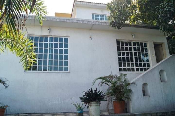A two-story house is shown, featuring large windows that allow natural light to enter. The exterior is painted in a light color, complemented by plants in terracotta pots. A staircase leads to the entryway, surrounded by greenery, enhancing the welcoming appearance.