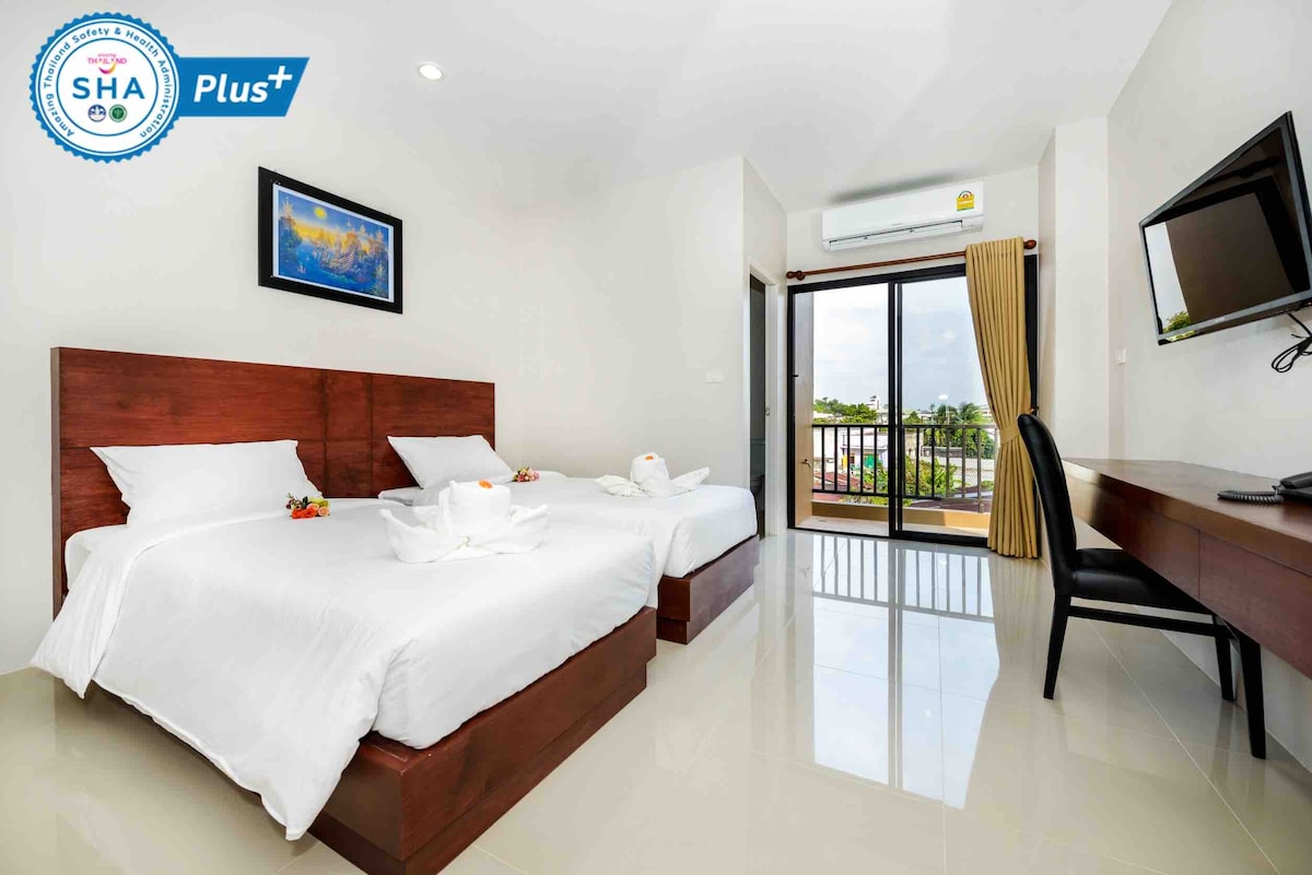 A well-lit hotel room features two neatly made beds with white linens, complemented by decorative towel arrangements. A desk and chair provide a workspace, while a glass door opens to a balcony, revealing views of the surroundings. Natural light brightens the space.