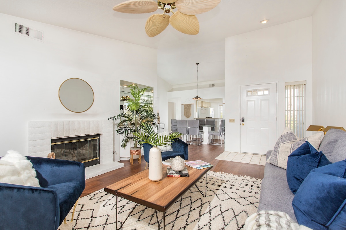 A living area features a comfortable seating arrangement with navy blue cushions. A wooden coffee table is set at the center, surrounded by a patterned area rug. A decorative plant and a circular wall mirror add visual interest, while a fireplace offers a cozy element.