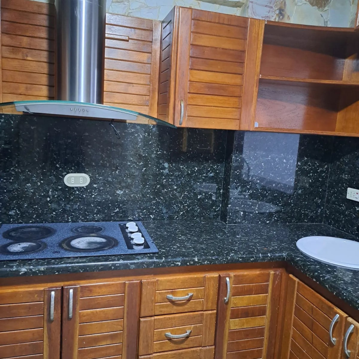 A modern kitchen features dark granite countertops and wooden cabinetry. A five-burner cooktop is positioned alongside a circular cutting board. The range hood overhead is metallic, and the backsplash is completed with matching granite for a cohesive look.