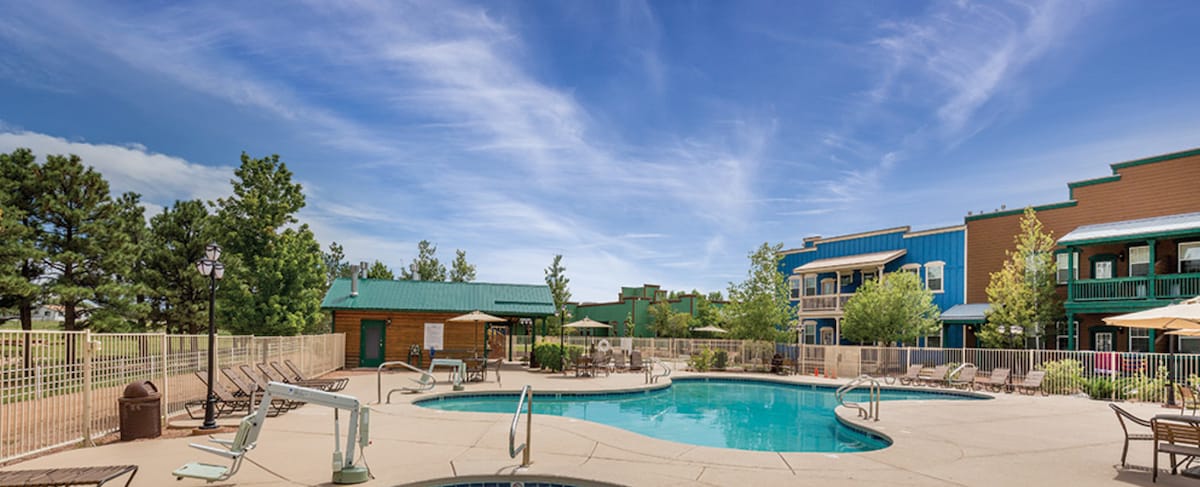 An outdoor pool area is framed by a secure fence, surrounded by shaded lounge chairs and umbrellas. Lush greenery and trees add to the serene atmosphere, while the blue sky enhances the inviting setting of the resort.