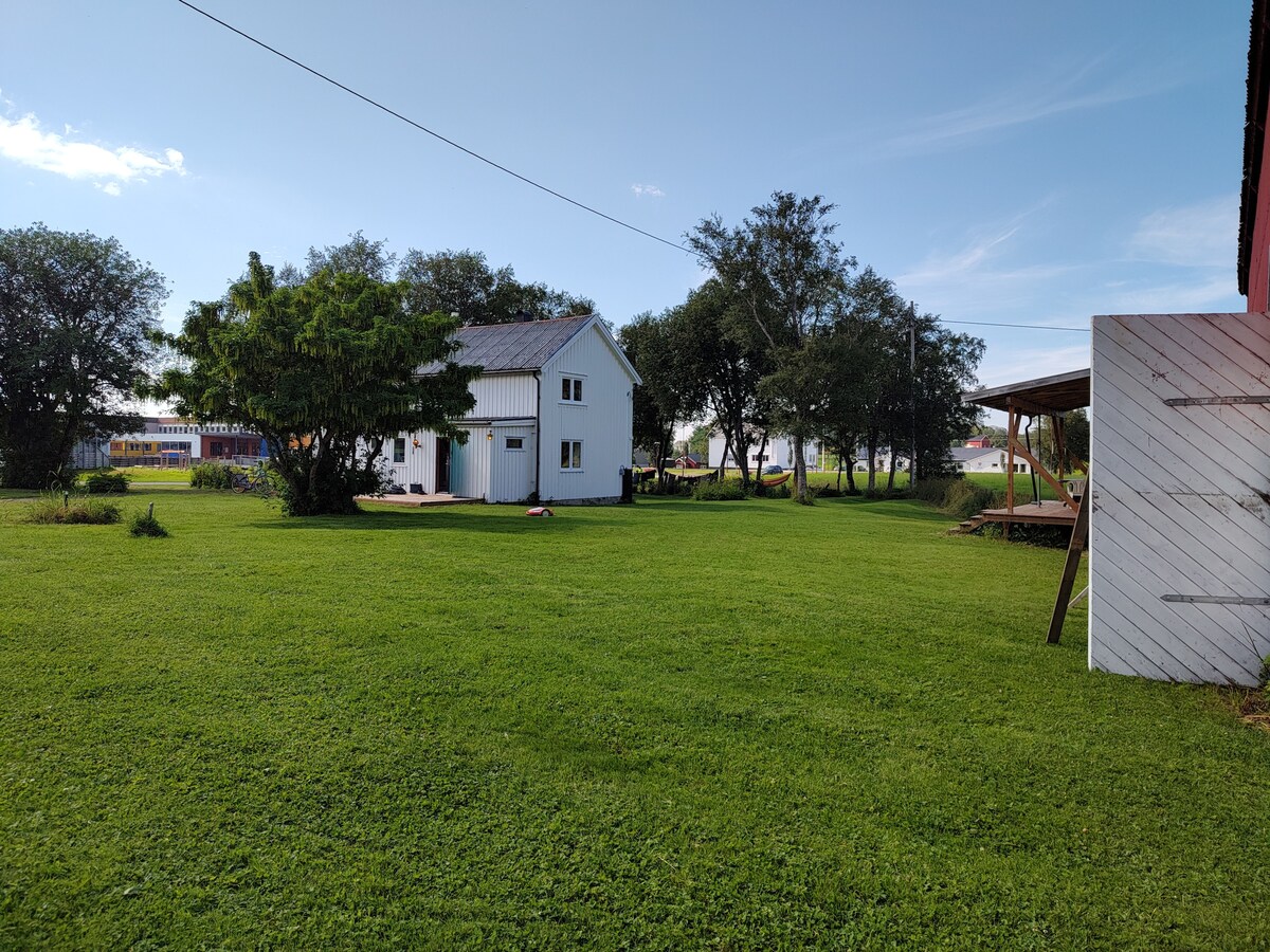 A well-maintained garden features a lush green lawn surrounded by trees. In the distance, a charming white house is visible, accompanied by a covered outdoor seating area. The serene setting is enhanced by the open space and clear blue sky.