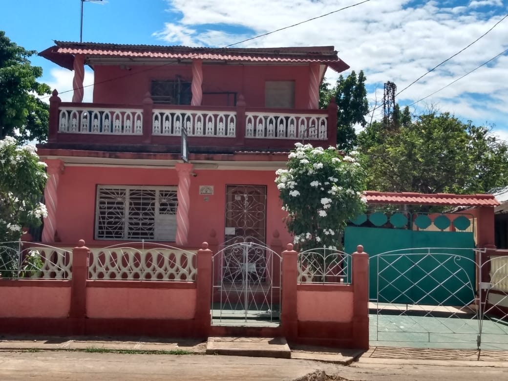 A two-story, pastel pink house is framed by lush greenery. The front yard features a decorative fence with floral accents. A balcony adorned with white railings overlooks the entrance, while the ground level includes large windows, creating a welcoming facade.