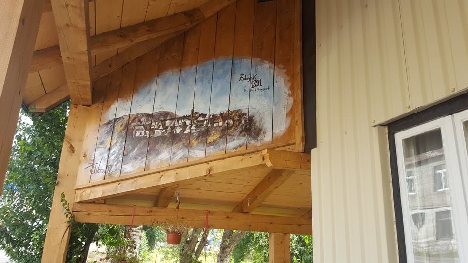 A wooden overhang features a painted mural depicting a scenic mountain village. The natural wooden beams and walls offer a rustic charm, complemented by greenery from surrounding trees. A window is visible on the right, providing additional light and connection to the outdoors.