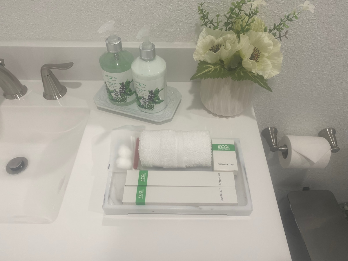 A clean bathroom countertop is displayed, featuring eco-friendly toiletries in green and white packaging. A white towel sits neatly beside cotton balls, and a decorative flower arrangement adds a touch of greenery. A toilet paper roll is visible, emphasizing the functional design.