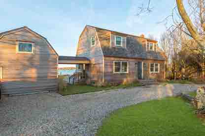 A charming two-story cottage is set against a backdrop of greenery. The light-colored wooden exterior is complemented by multiple windows, bringing in natural light. A gravel driveway leads to the entrance, with a landscaped yard enhancing the overall appeal.