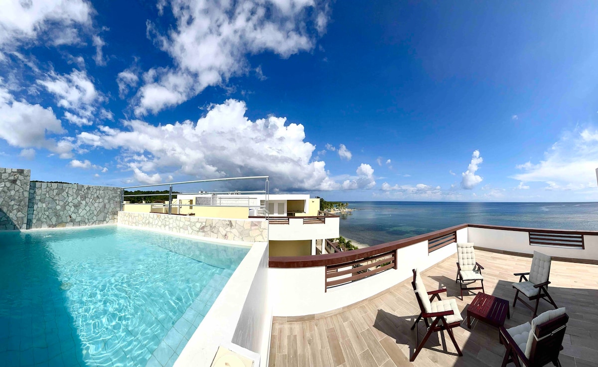 A spacious terrace overlooks the sea, featuring a private pool with clear blue water. Two lounge chairs and a small table are positioned to enjoy the panoramic views of the horizon and sky, illuminated by soft sunlight and scattered clouds.