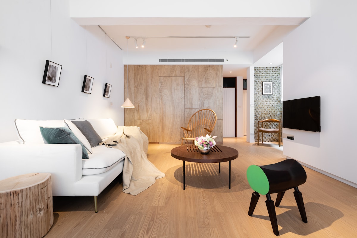 The spacious living area features a comfortable sofa adorned with cushions, a round coffee table at its center, and a variety of wall art. Light wood flooring complements the natural elements, while a decorative chair adds vibrant color and interest to the room.