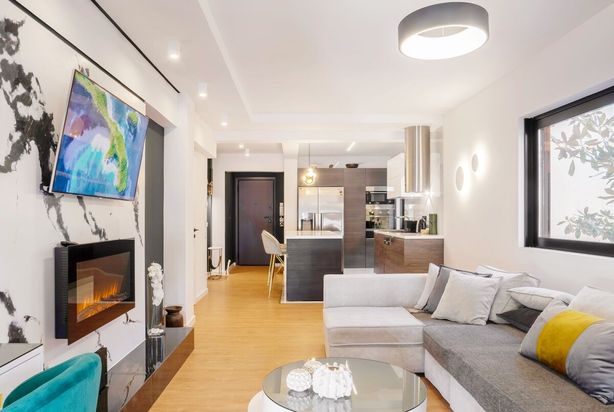 A modern living area features a sleek marble wall with a mounted television and a cozy electric fireplace. A spacious sectional sofa with various cushions faces a glass coffee table. The open layout extends towards a contemporary kitchen, enhancing accessibility and comfort.
