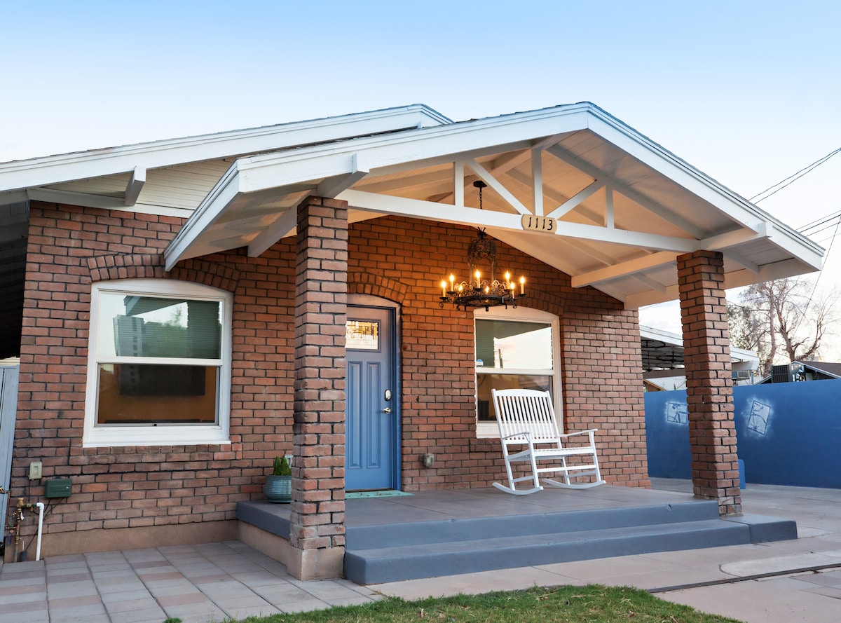 A historic brick bungalow showcases a covered porch, featuring a white rocking chair and a charming blue front door. Two large windows allow natural light to enter, and a chandelier hangs from the ceiling, adding unique character to the entrance.