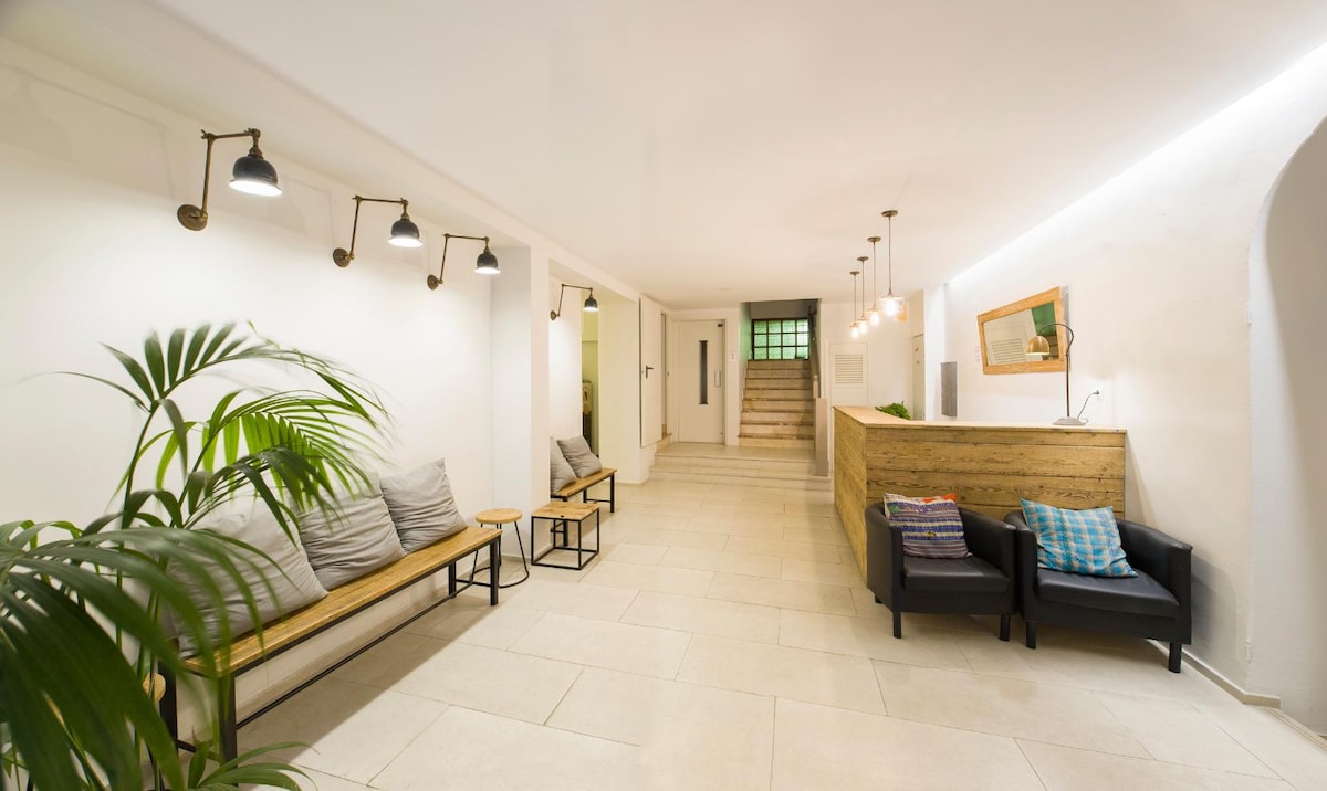 A bright reception area features modern seating arrangements with benches and armchairs. Natural light enters through a nearby window, illuminating light-colored walls and tiled flooring. Decorative elements, including plants and wall-mounted lighting fixtures, contribute to a welcoming environment.