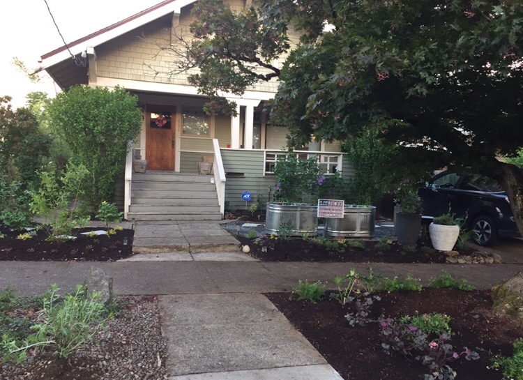 The exterior features a welcoming Craftsman-style house with a wooden front door and steps leading to the entrance. Well-maintained gardens with various plants and decorative pots flank either side of the pathway, creating an inviting approach to the home.
