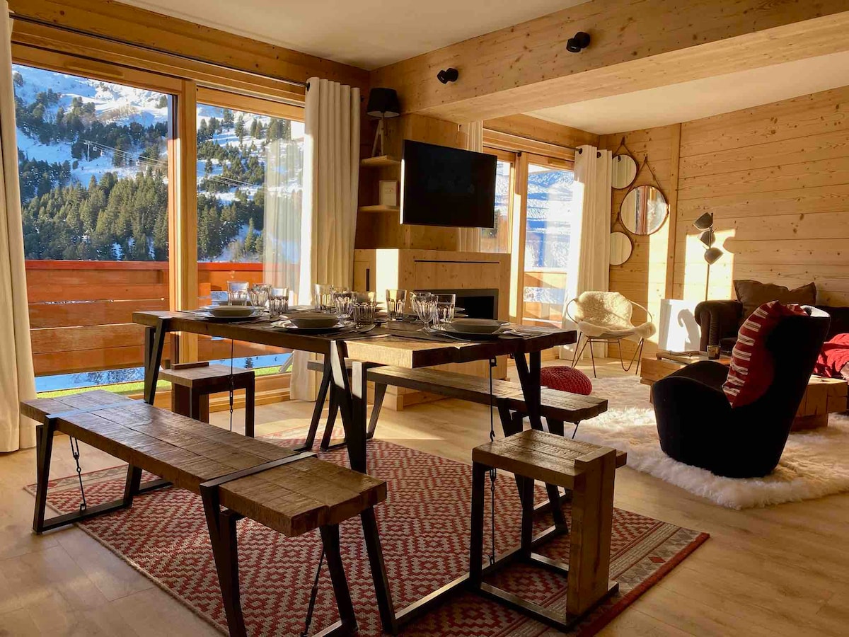 A warm living area features large glass windows offering panoramic views of the mountains. A dining table with benches is set for meals, and a comfortable black chair sits nearby. Natural wood walls and a plush rug contribute to a cozy, inviting atmosphere.