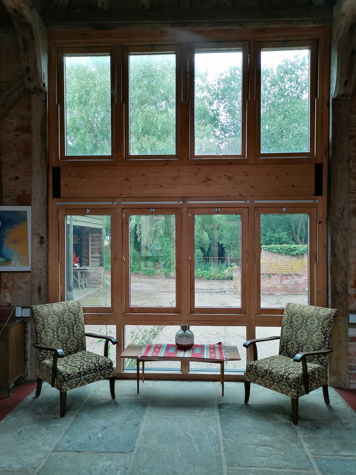 Two patterned chairs are positioned beside a small wooden table, facing large windows that offer views of the garden. Natural light enters the space, illuminating the stone flooring and highlighting the rustic wooden beams of the barn.