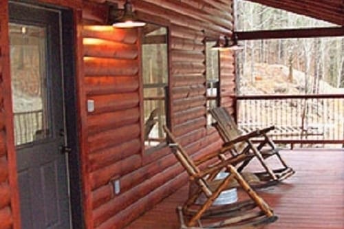 A covered deck area features two wooden rocking chairs positioned invitingly. The log cabin exterior is highlighted by natural wood tones, with large windows allowing visibility of the natural surroundings. The entrance door is situated beside the deck, blending seamlessly with the cabin's rustic design.