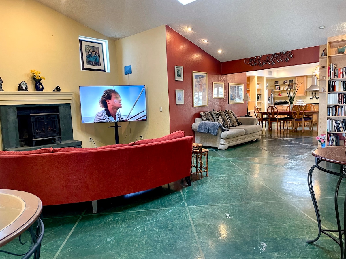 A spacious living area features a large television mounted on a stand and a fireplace against a colorful wall. Seating includes a cozy sofa and a curved red couch. Light reflects off the green tiled floor, connecting seamlessly to the dining area in the background.